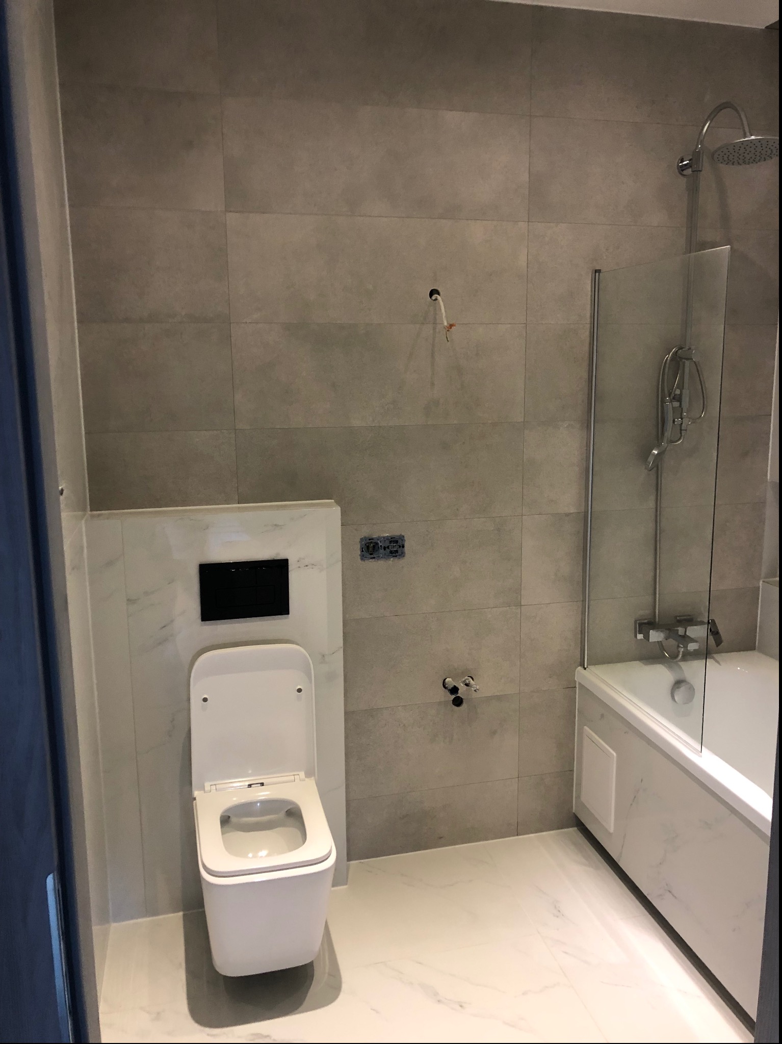 Nowoczesna, wykończona łazienka z szarymi płytkami, białą armaturą, kabiną prysznicową ze szklaną ścianką i podtynkową spłuczką toaletową, widoczne niedokończone przyłącza wodne.