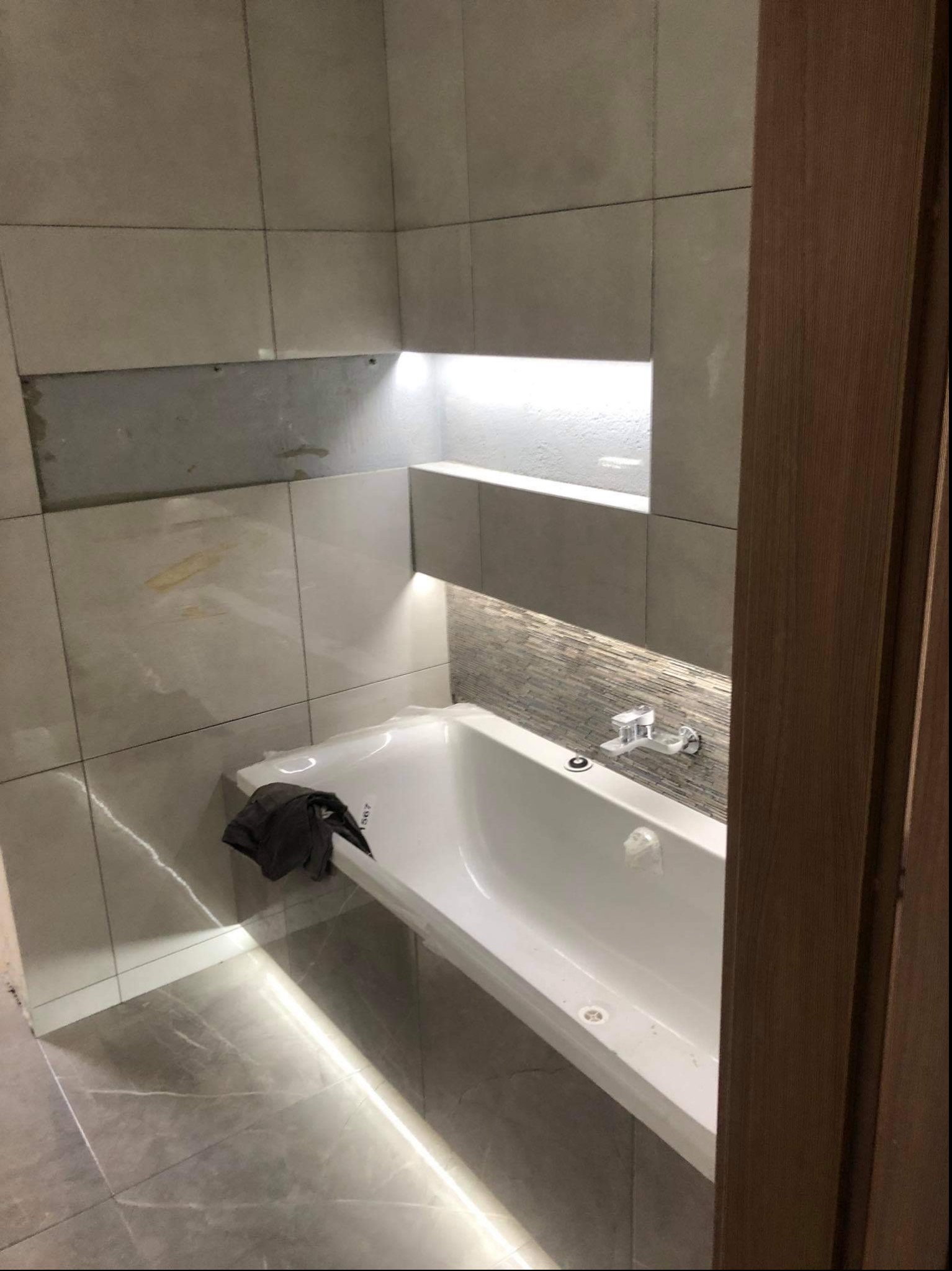 Łazienka w trakcie wykańczania: biała wanna z akrylowym zabezpieczeniem, szara płytka imitująca kamień na ścianach, podświetlane wnęki, brakująca płytka nad wanną.