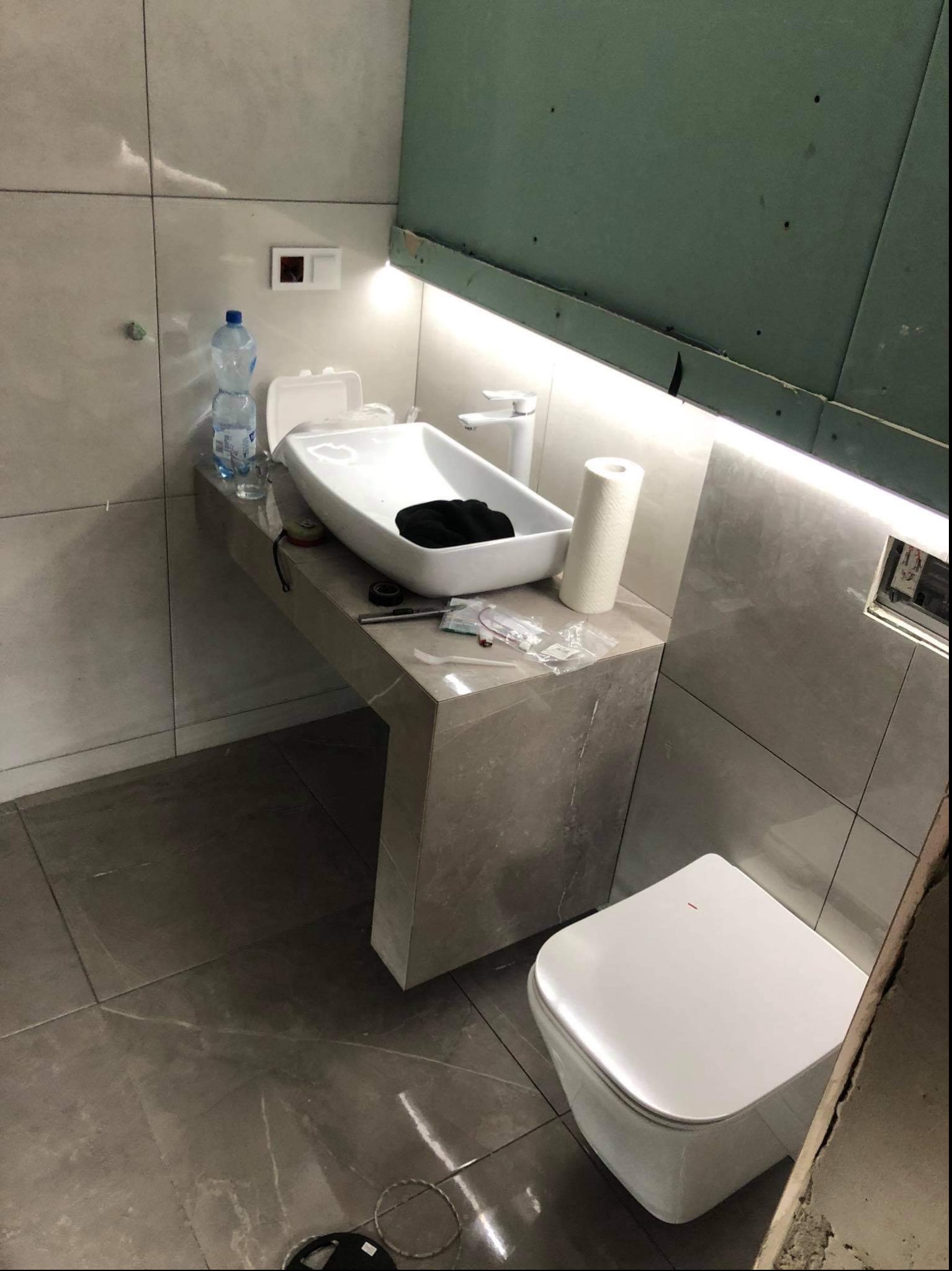 Łazienka w trakcie prac wykończeniowych: podwieszana umywalka na blacie z imitacji kamienia, wisząca toaleta, płytki imitujące kamień na ścianach i podłodze, widoczne narzędzia i materiały...