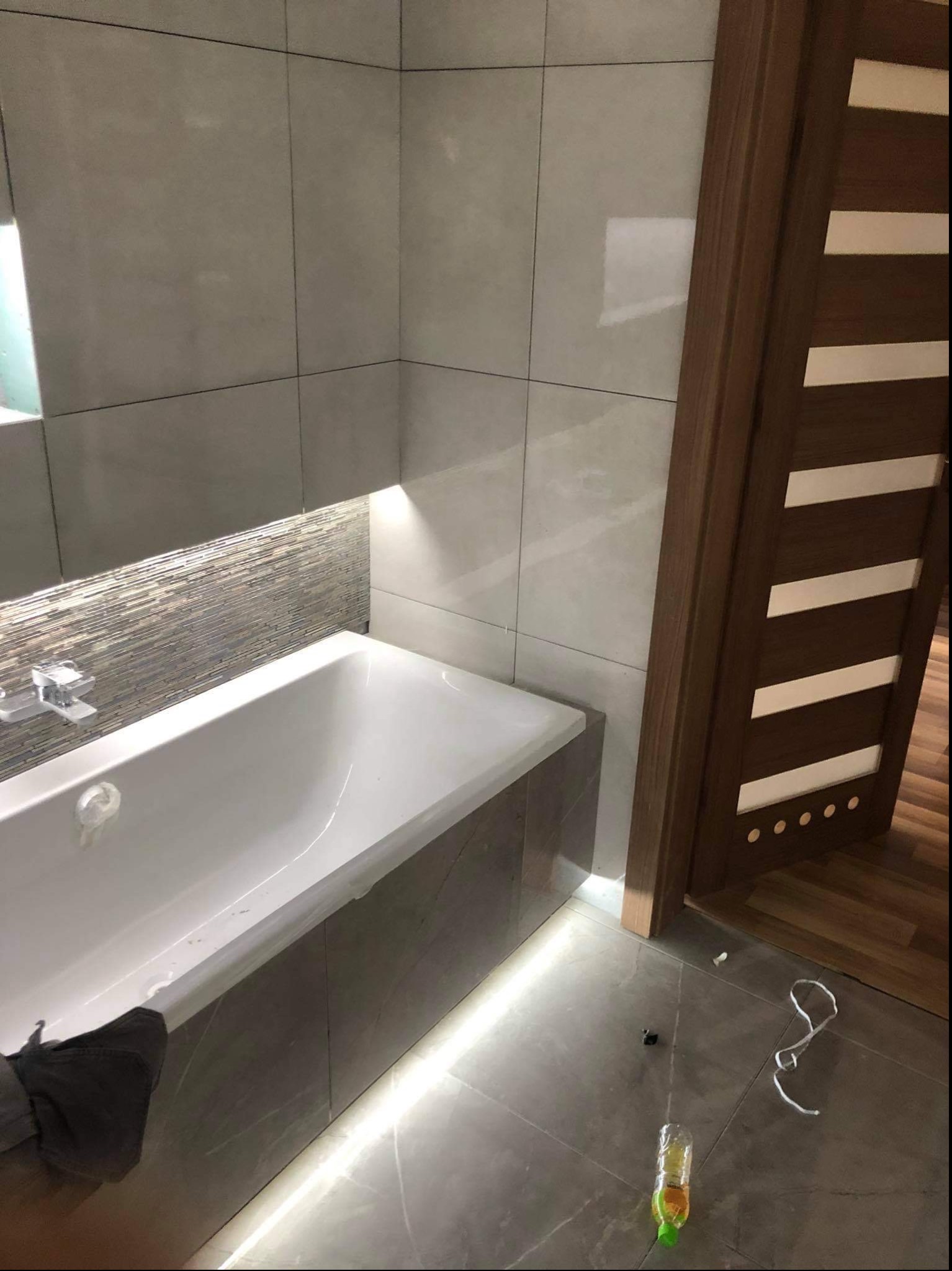 Wykończona łazienka z wanną obudowaną płytkami w odcieniach szarości, mozaika nad wanną z podświetleniem LED, szare płytki na ścianach i podłodze, widoczne niedociągnięcia na podłodze (śmieci, kabel)