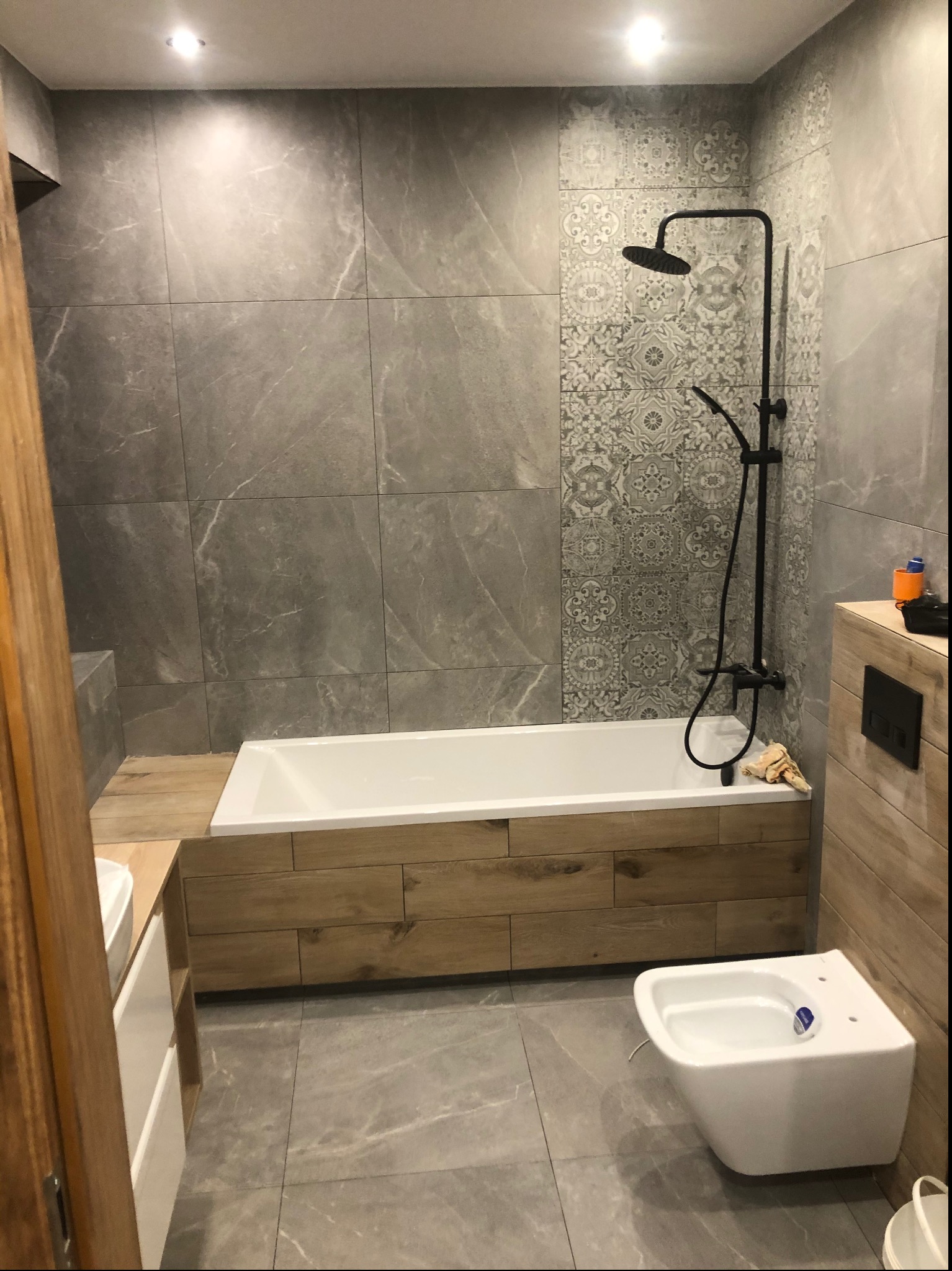 Nowoczesna łazienka z szarymi płytkami imitującymi marmur, dekoracyjnym pasem płytek wzorzystych, białą wanną obudowaną drewnopodobnymi panelami, czarną armaturą natryskową i podwieszanym bidetem.