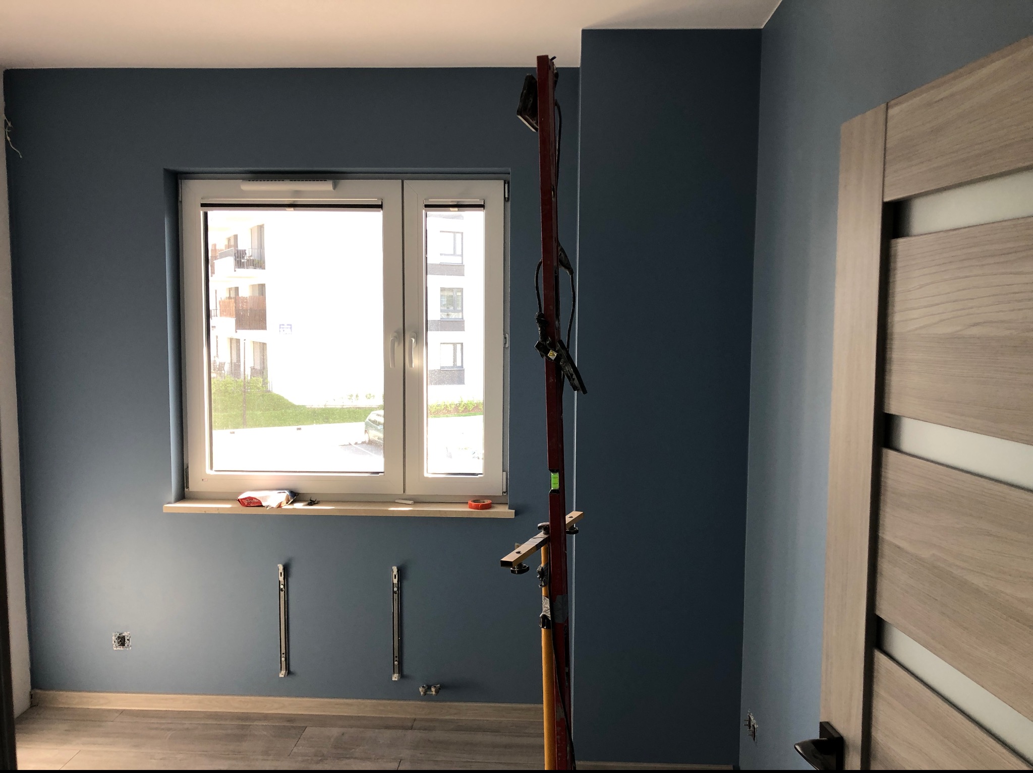 Świeżo pomalowane na niebiesko ściany pokoju z zamontowanym oknem i grzejnikiem, widoczna drabina malarska i fragment drewnianych drzwi.