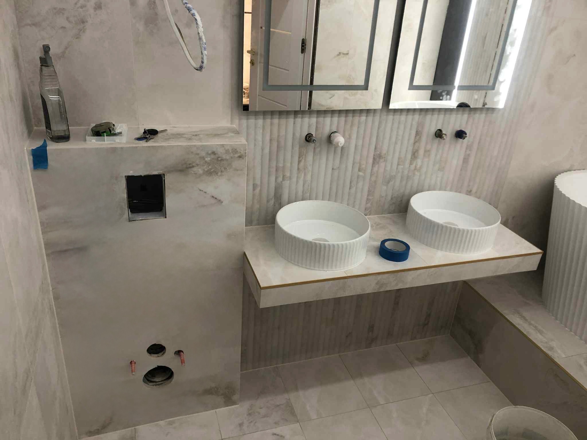 Łazienka w trakcie wykańczania: dwie białe umywalki na blacie z płytek, lustra z podświetleniem LED, widoczne instalacje i narzędzia. Wnętrze w odcieniach szarości i bieli.