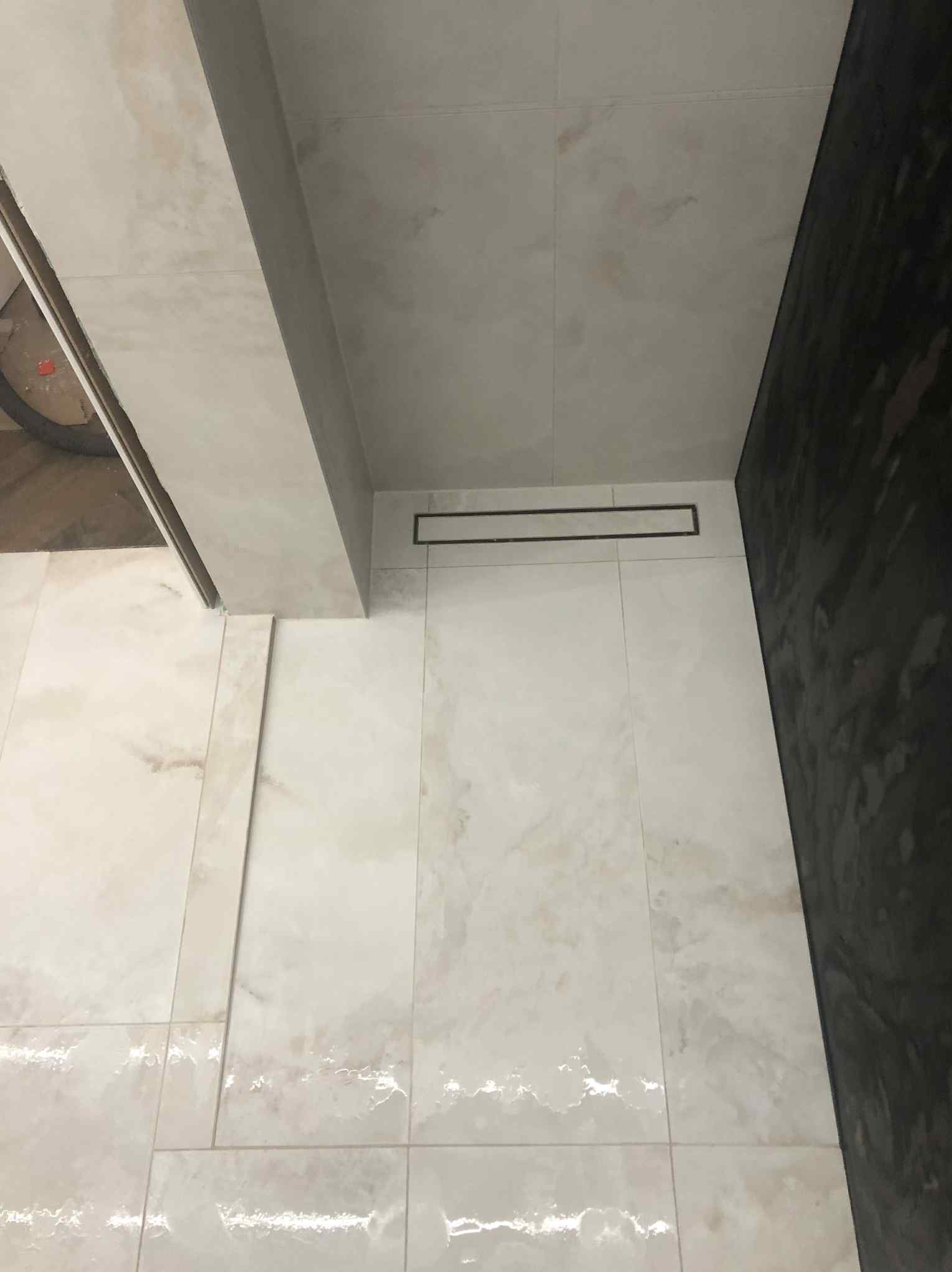 Wykończona łazienka z beżowymi płytkami imitującymi marmur i odpływem liniowym. Ciemna ściana w kabinie prysznicowej. Widok z góry.