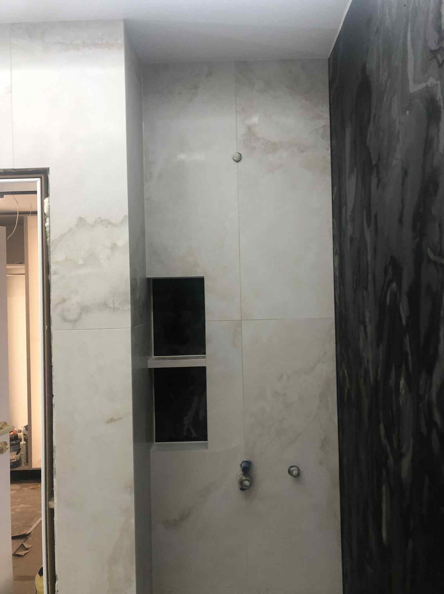 Wykończona ściana łazienki z jasnymi płytkami imitującymi marmur, wnęką z czarnymi półkami i fragmentem ściany pokrytej ciemnym tynkiem dekoracyjnym.