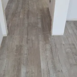 Długi korytarz z świeżo położoną, jasną podłogą imitującą drewno, widoczne surowe ściany i niedokończone wykończenia.