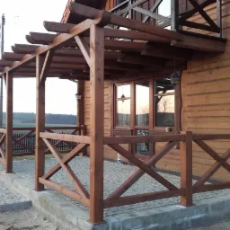 Drewniana pergola przylegająca do domu z bali, z widokiem na polanę w tle. Konstrukcja z krzyżowymi elementami, oświetlona wiszącymi lampami.