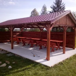 Zadaszona konstrukcja z drewnianymi słupami i ścianami, kryta bordową blachodachówką, z rzędami drewnianych stołów i ławek, na betonowej wylewce, otoczona zielonym trawnikiem.