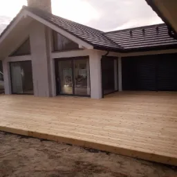 Nowy taras z jasnego drewna przed nowoczesnym domem z dużymi oknami przesuwnymi, widoczny samochód dostawczy i narzędzie na piasku.