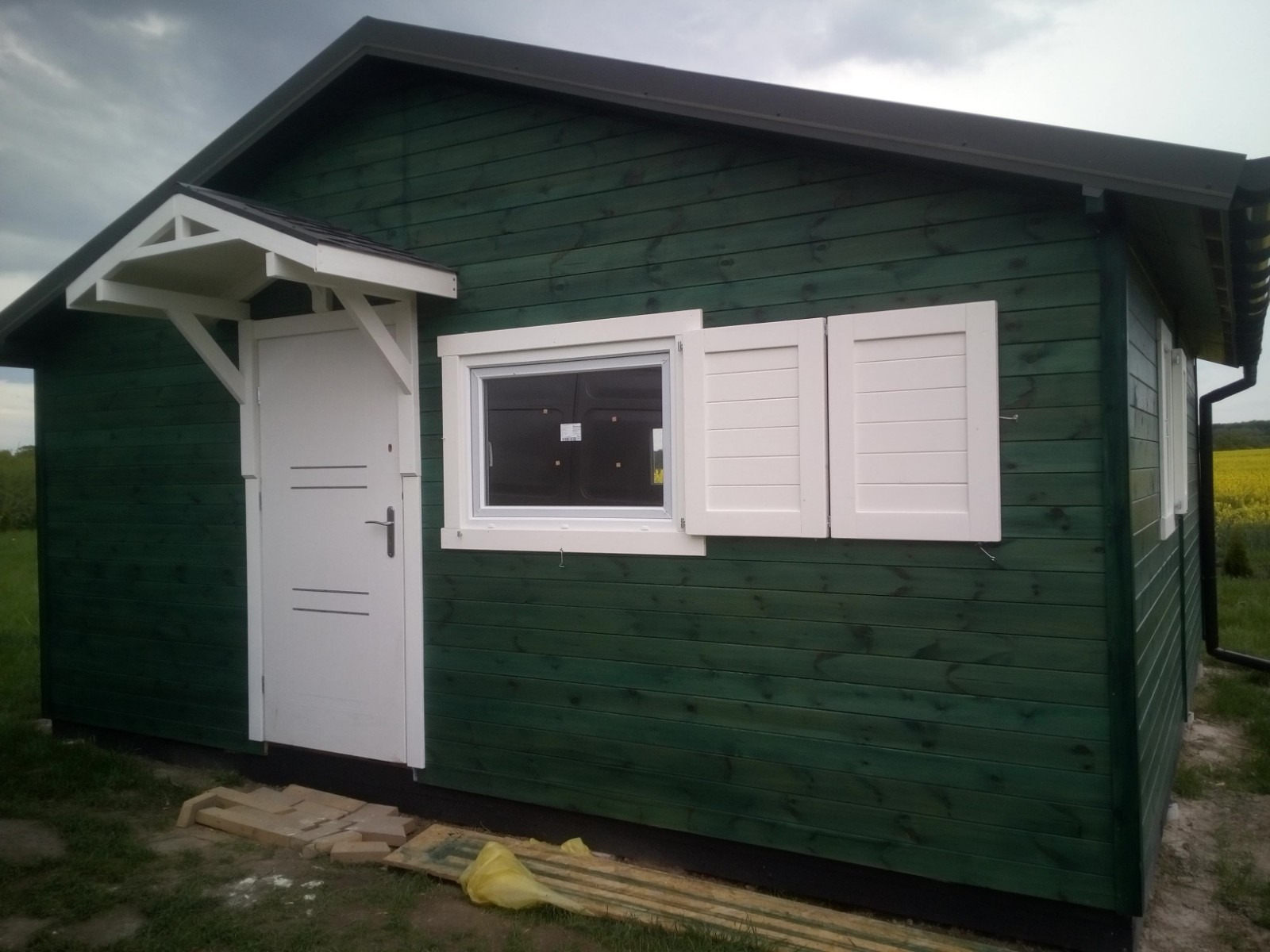 Zielony domek letniskowy z białymi elementami wykończeniowymi: okna z okiennicami, drzwi z zadaszeniem. Widoczne żółte pole w tle.