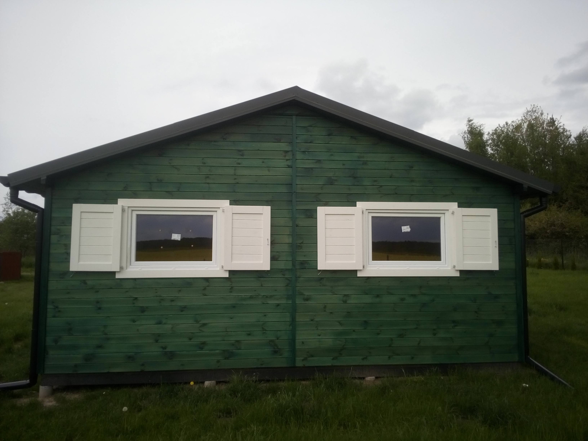 Zielony domek letniskowy z drewnianą elewacją i białymi okiennicami, na tle zielonej trawy i pochmurnego nieba.