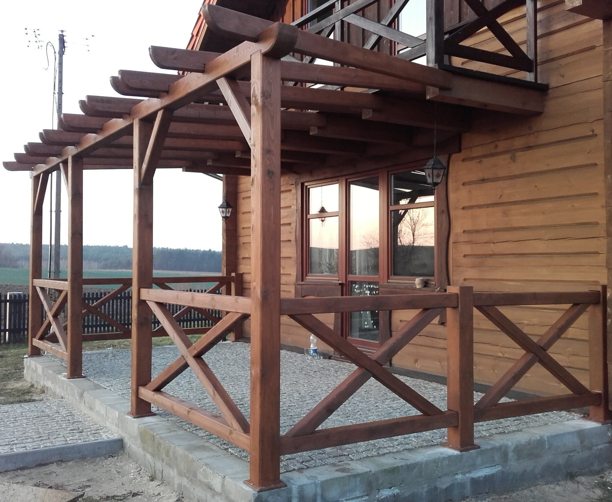 Drewniana pergola przylegająca do domu z bali, z widokiem na polanę w tle. Konstrukcja z krzyżowymi elementami, oświetlona wiszącymi lampami.