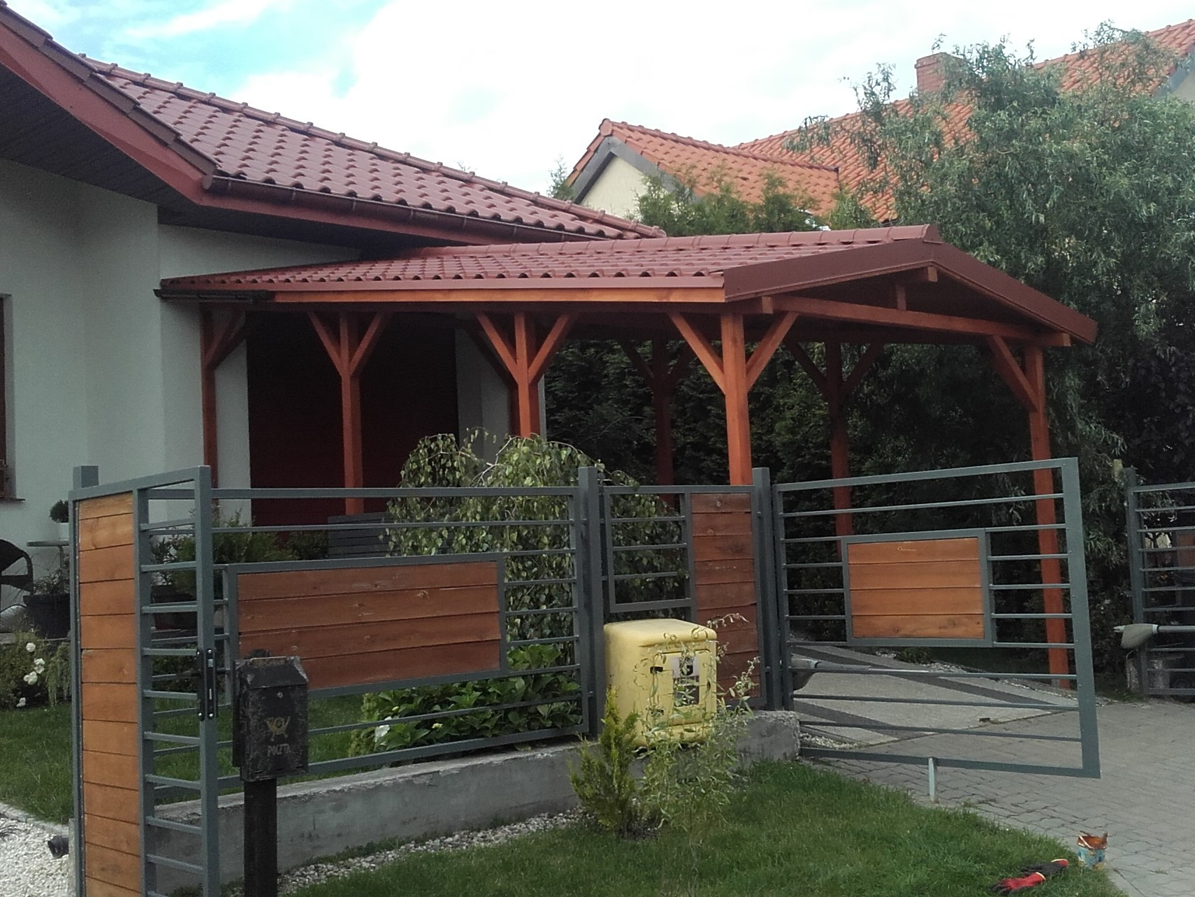 Zadaszenie w kolorze brązowym, wsparte na drewnianych słupach, przylegające do jasnego domu z czerwoną dachówką, widoczne zza metalowo-drewnianego ogrodzenia z otwartą bramą i żółtą skrzynką pocztową.
