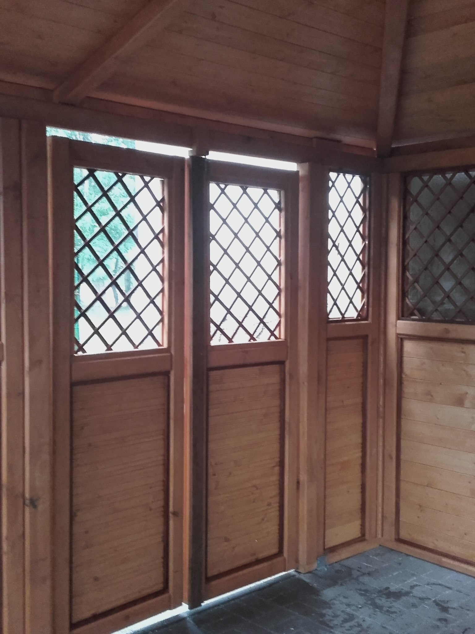 Wnętrze drewnianej konstrukcji z ażurowymi oknami, widoczne elementy konstrukcyjne i podłoga, częściowo przesłonięte światłem z zewnątrz.