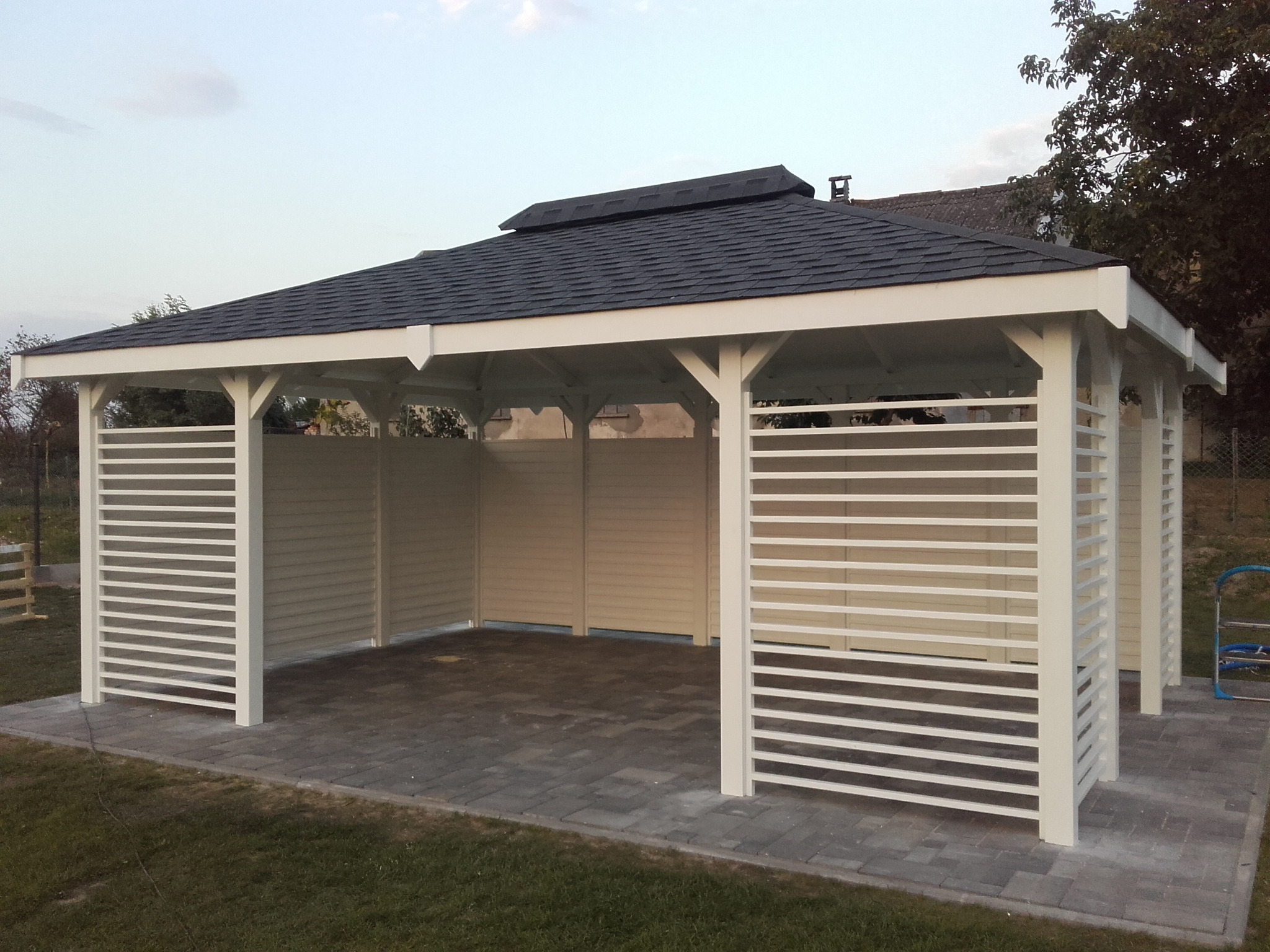 Biała altana ogrodowa z czarnym dachem z gontu bitumicznego, częściowo osłonięta poziomymi listwami, na wybrukowanym placu, widok z zewnątrz na tle zieleni i budynków.