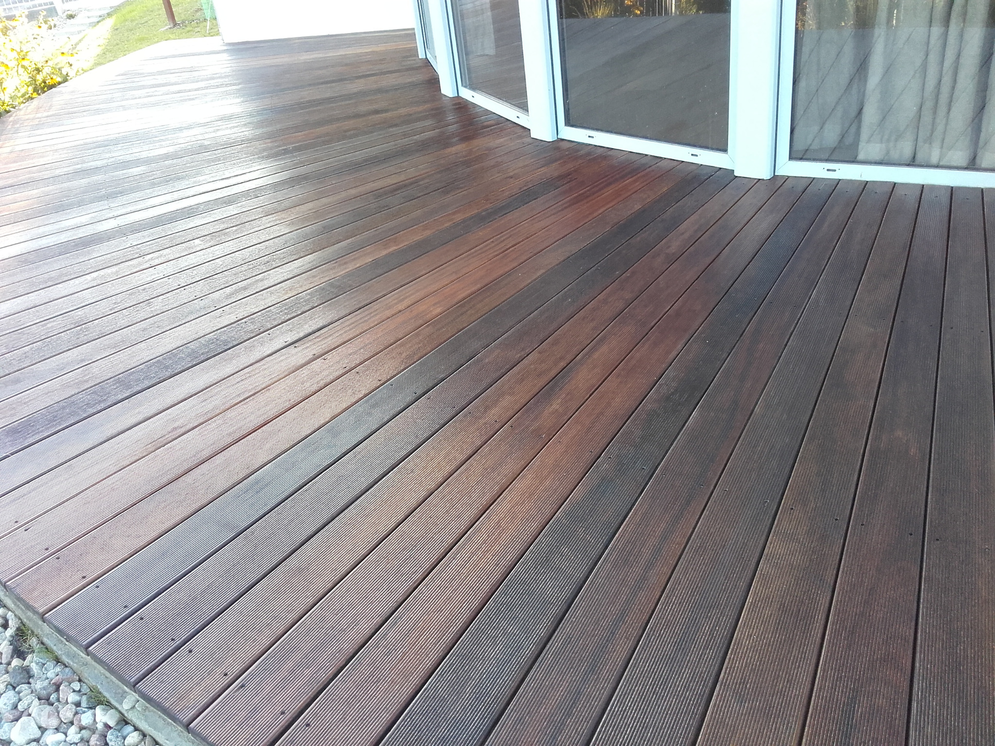 Nowoczesny taras z desek kompozytowych w odcieniach brązu, przylegający do budynku z dużymi przesuwnymi oknami, wykończony drobnymi kamieniami na obrzeżach.