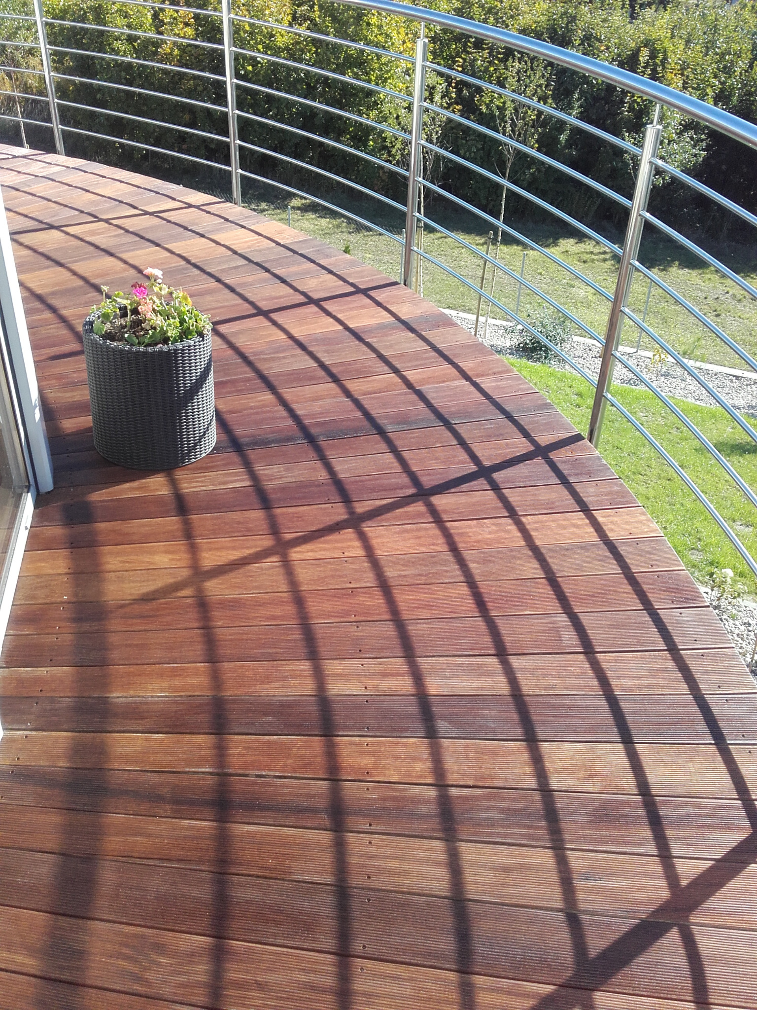 Zakrzywiony taras z desek w kolorze ciepłego brązu, z metalową balustradą i donicą z kwiatami, oświetlony słońcem rzucającym cienie balustrady.