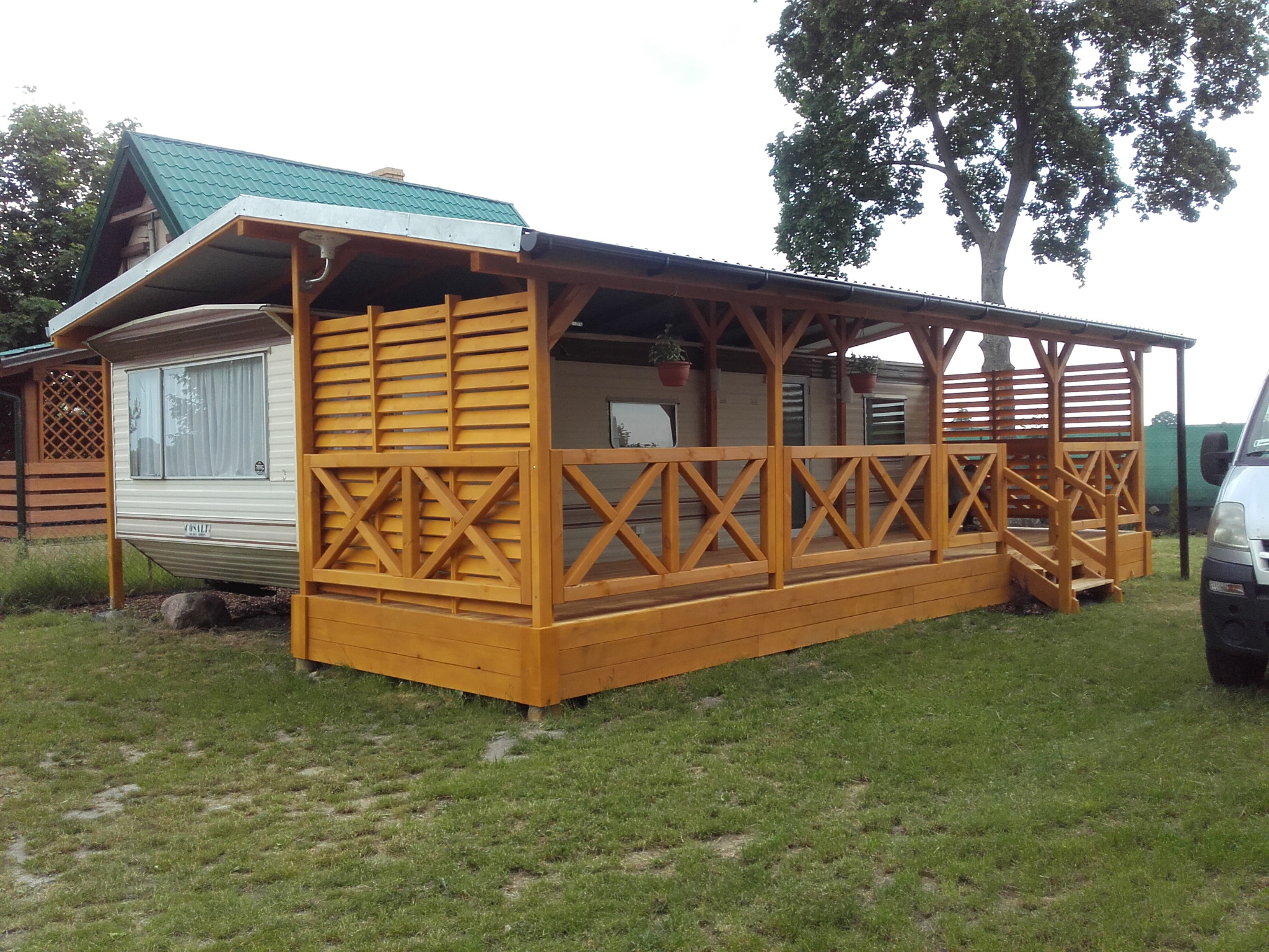 Drewniana altana z zadaszeniem przylegająca do przyczepy kempingowej w Grudziądzu, z dekoracyjnymi elementami krzyżowymi i doniczkami z kwiatami, widok z poziomu gruntu.