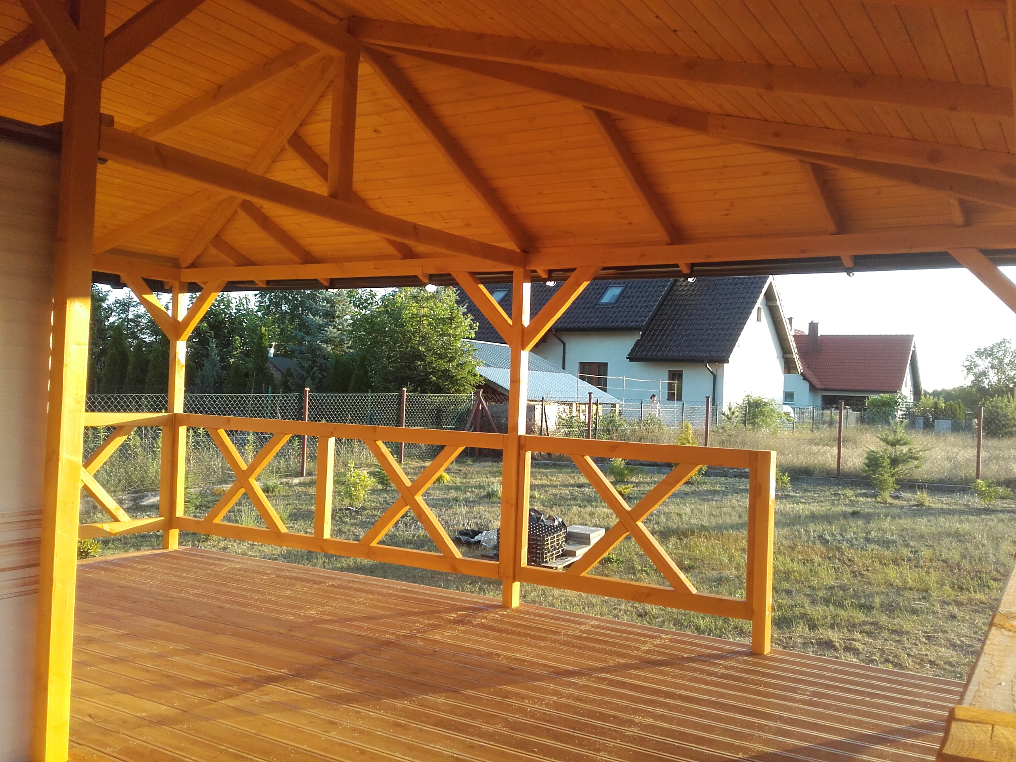 Zadaszona konstrukcja z drewnianym dachem i balustradą w kształcie krzyży, widok na ogród i zabudowania w tle.