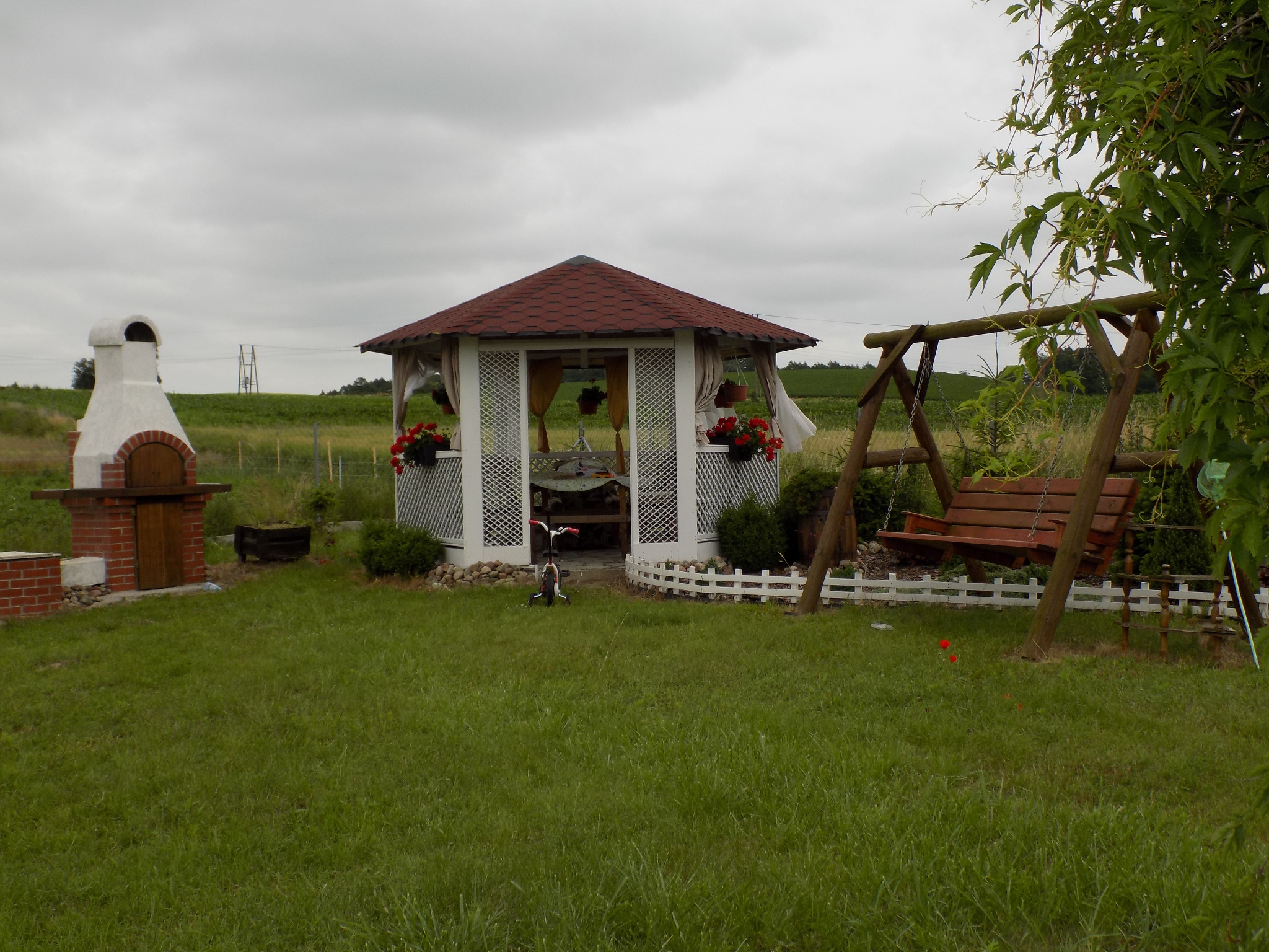 Ogród z białą, ośmiokątną konstrukcją z czerwonym dachem, kwiatami w donicach, huśtawką i murowanym grillem.