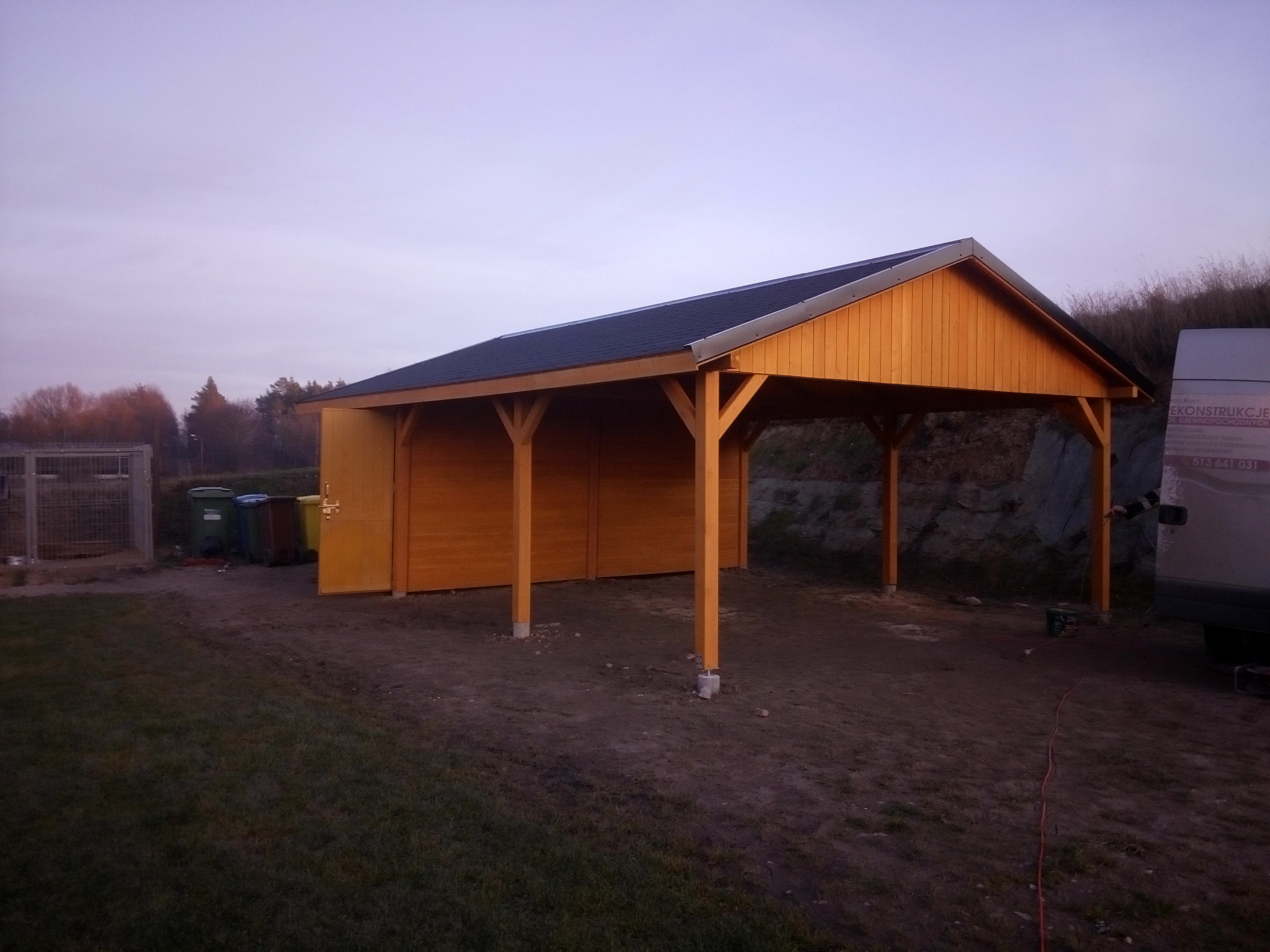 Drewniana konstrukcja z zadaszeniem, prawdopodobnie wiata garażowa lub altana z pomieszczeniem gospodarczym z boku, widoczna na tle zachodzącego słońca, obok zaparkowany samochód dostawczy.
