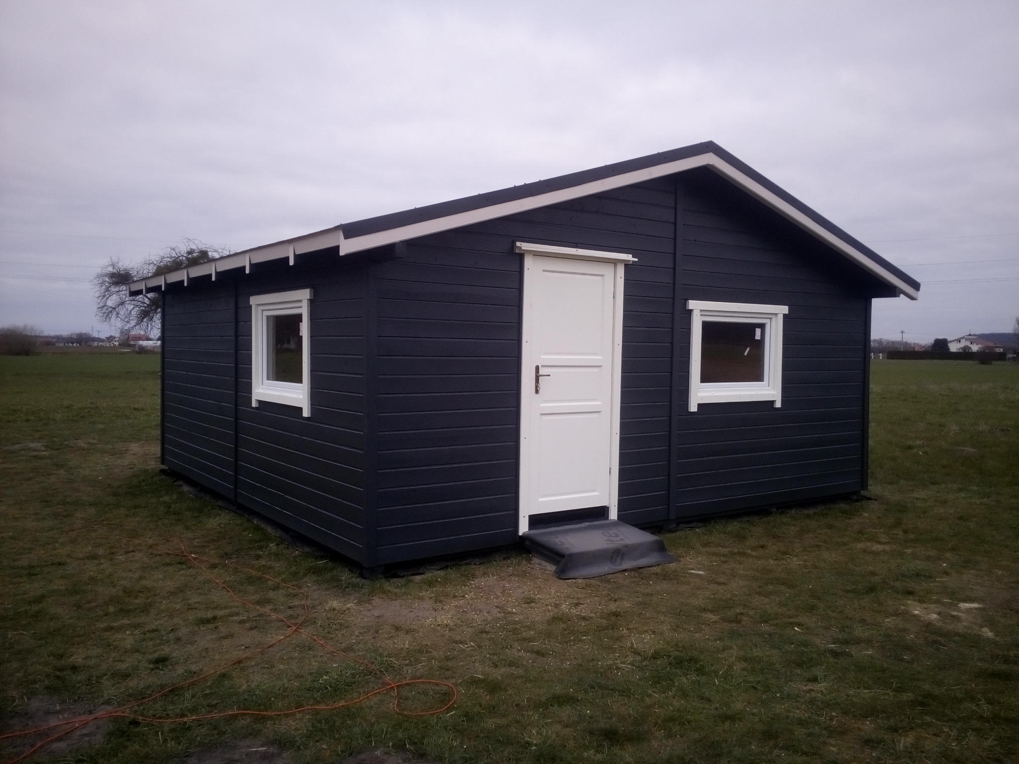 Gotowa, ciemna, drewniana konstrukcja z białymi obramowaniami okien i drzwi, umieszczona na trawiastym terenie pod pochmurnym niebem.