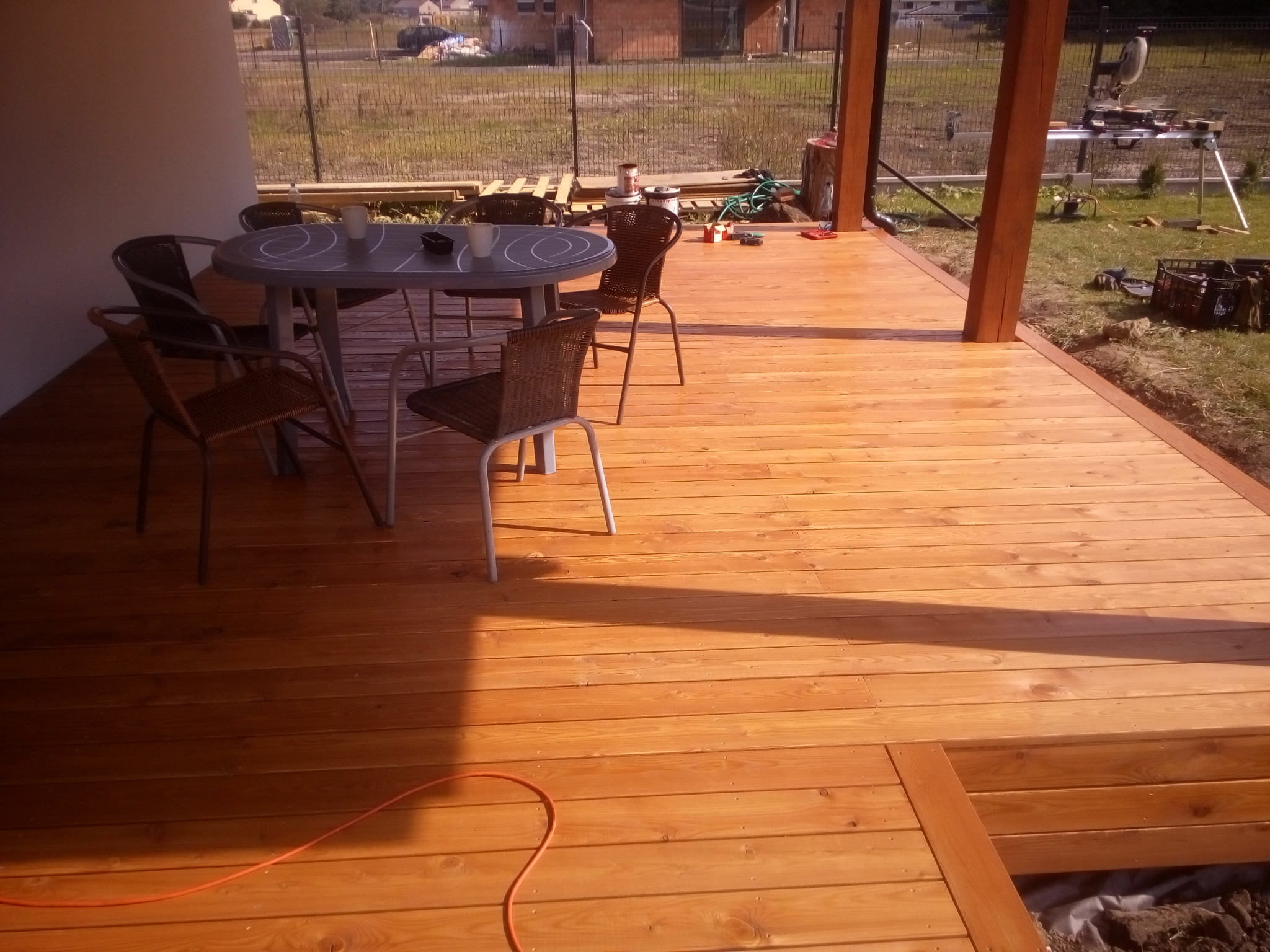 Świeżo wybudowany taras z drewna, z widocznymi narzędziami pracy na trawie w tle, z szarym okrągłym stołem i krzesłami z wikliny na nim.