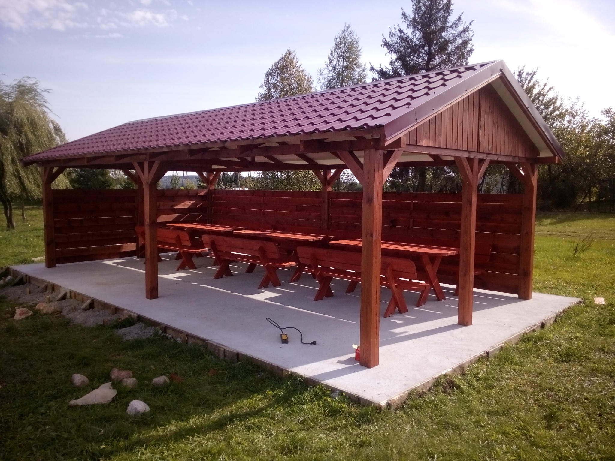 Zadaszona konstrukcja z drewnianymi słupami i ścianami, kryta bordową blachodachówką, z rzędami drewnianych stołów i ławek, na betonowej wylewce, otoczona zielonym trawnikiem.