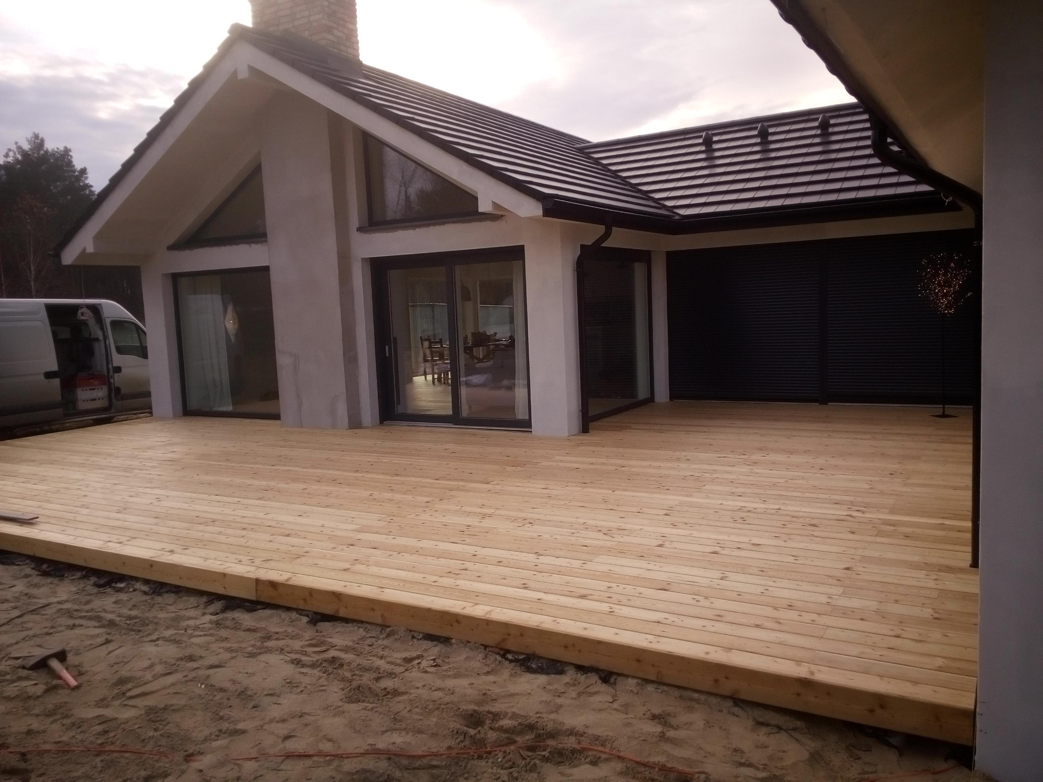 Nowy taras z jasnego drewna przed nowoczesnym domem z dużymi oknami przesuwnymi, widoczny samochód dostawczy i narzędzie na piasku.