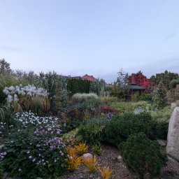 BUXUS Wiktor Skotarczak - Bujny, wielobarwny ogród z kwitnącymi roślinami, ozdobnymi trawami i krzewami, otoczony kamieniami. W tle widoczny domek ogrodowy i zabudowania.
