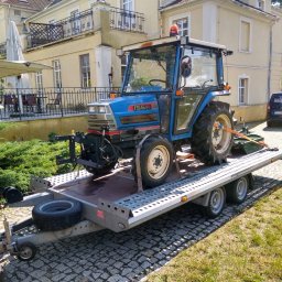 BUXUS Wiktor Skotarczak - Niebieski traktor Iseki na przyczepie transportowej na tle budynku z balkonem i zaparkowanego auta. Widoczny fragment osoby. Kostka brukowa i trawa.