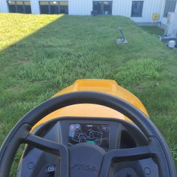 BUXUS Wiktor Skotarczak - Widok z perspektywy operatora na żółty traktorek Stiga podczas koszenia trawy przed budynkiem. Nierówna trawa, cień na trawniku.