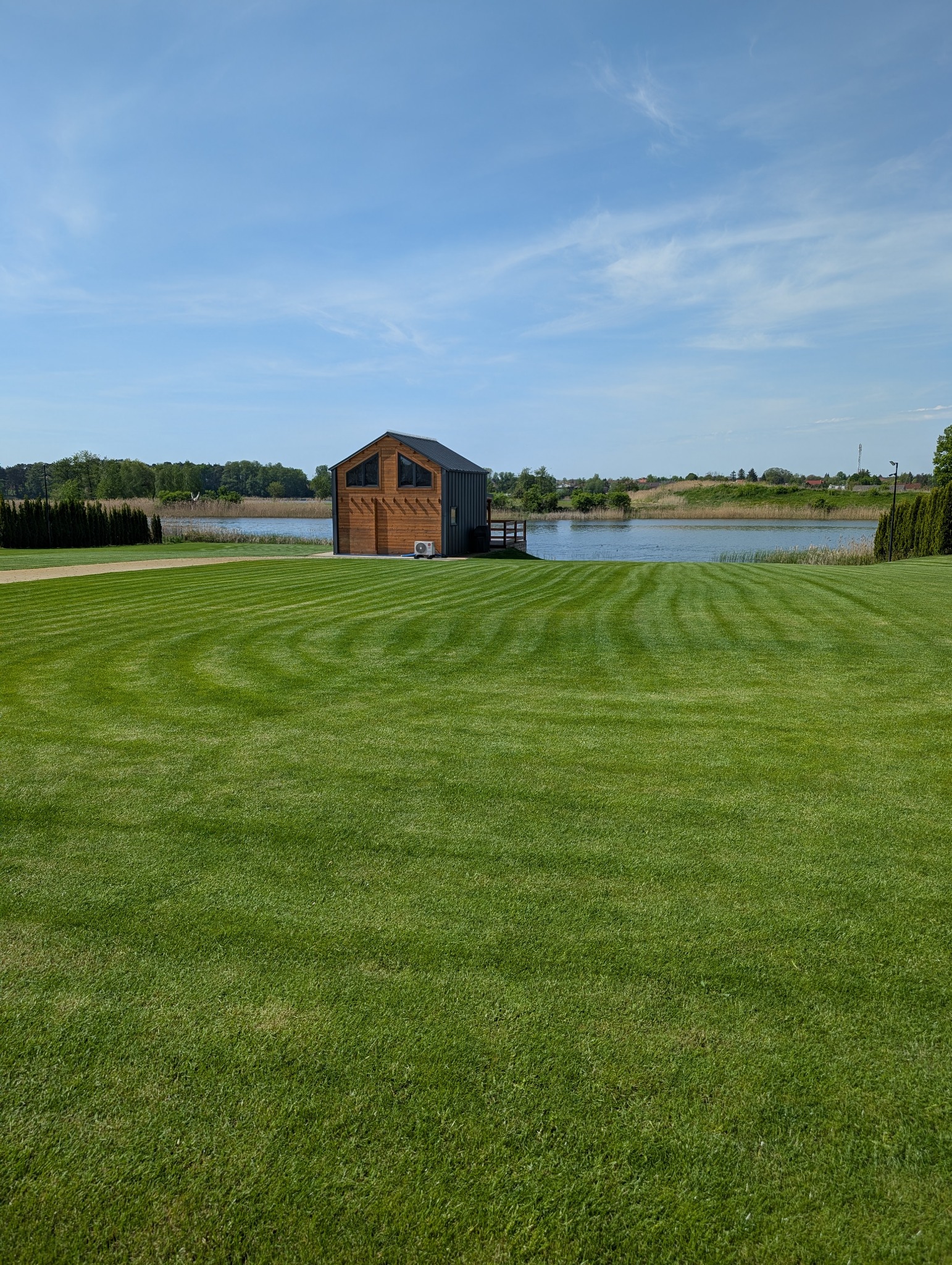 Perfekcyjnie skoszony trawnik z geometrycznym wzorem, prowadzący do drewnianego domku nad jeziorem, w słoneczny dzień. Widoczne pasy koszenia.