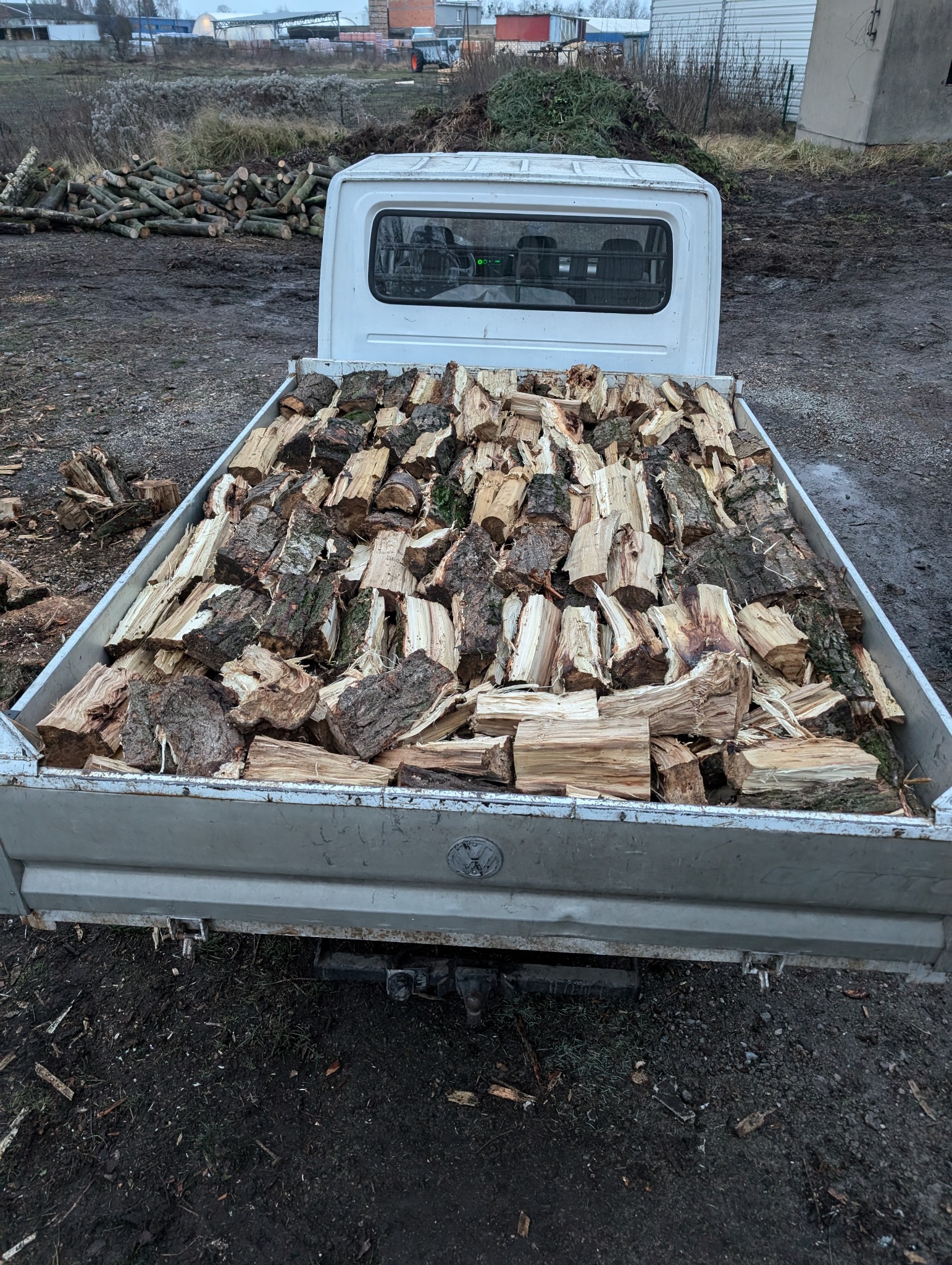 Ciężarówka wypełniona pociętym drewnem na tle sterty gałęzi i pni. Widoczne błoto i resztki drewna na ziemi. Szara kolorystyka, pochmurny dzień.