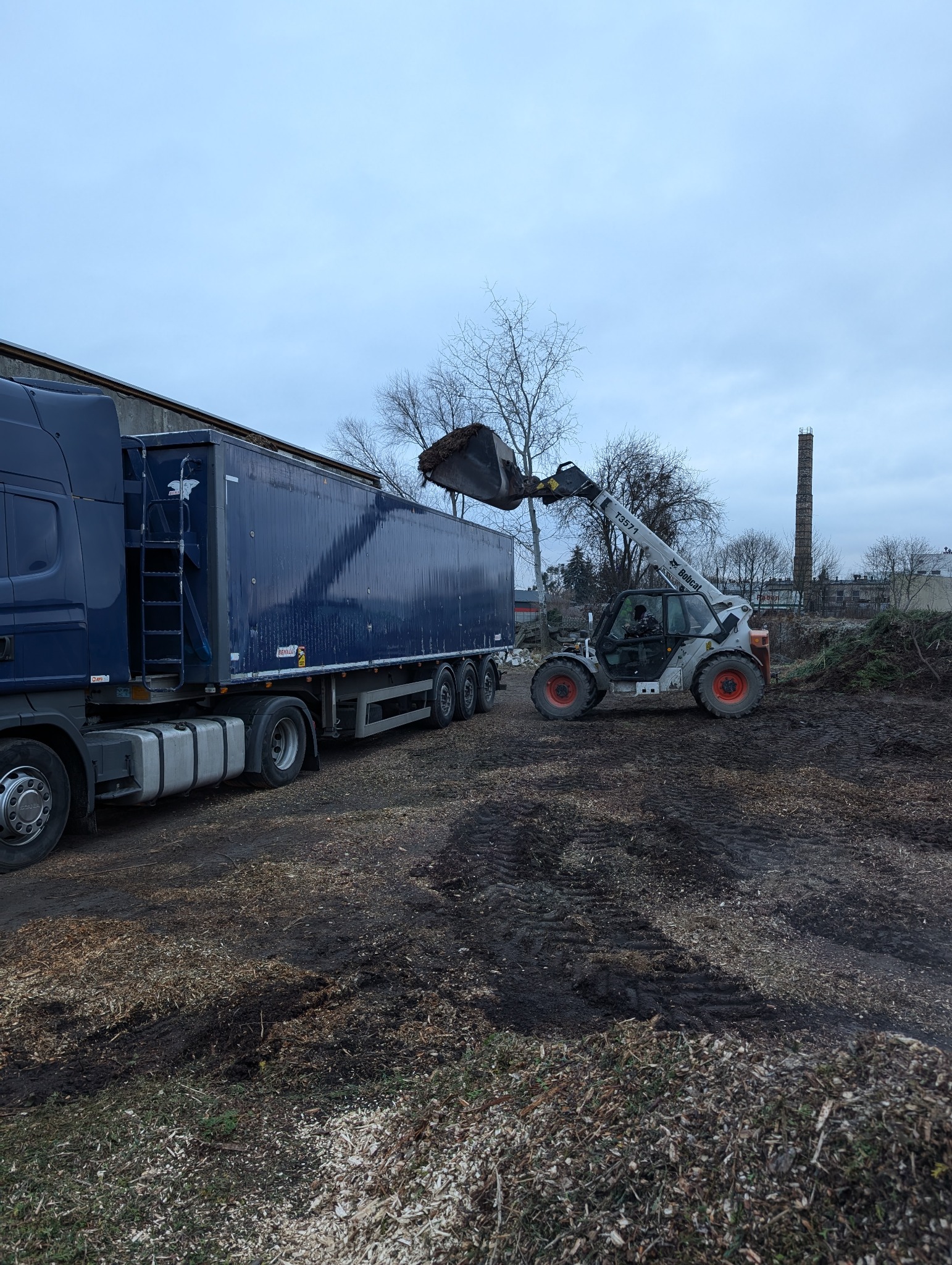 Bobcat T3571 ładuje rozdrobnione gałęzie do naczepy ciężarówki na tle pochmurnego nieba i komina w oddali. Widoczne ślady opon na ziemi.