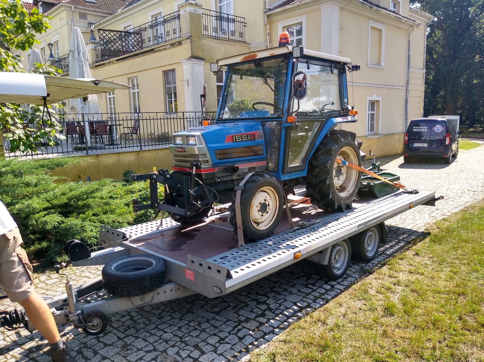 Niebieski traktor Iseki na przyczepie transportowej na tle budynku z balkonem i zaparkowanego auta. Widoczny fragment osoby. Kostka brukowa i trawa.