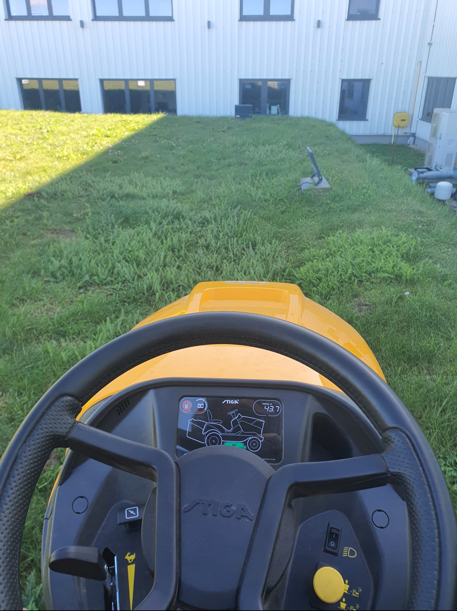 Widok z perspektywy operatora na żółty traktorek Stiga podczas koszenia trawy przed budynkiem. Nierówna trawa, cień na trawniku.