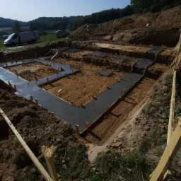 Wylane fundamenty pod budowę domu, widoczny szalunek i świeży beton, teren przygotowany pod dalsze prace budowlane, w tle krajobraz zalesiony i zaparkowane samochody.