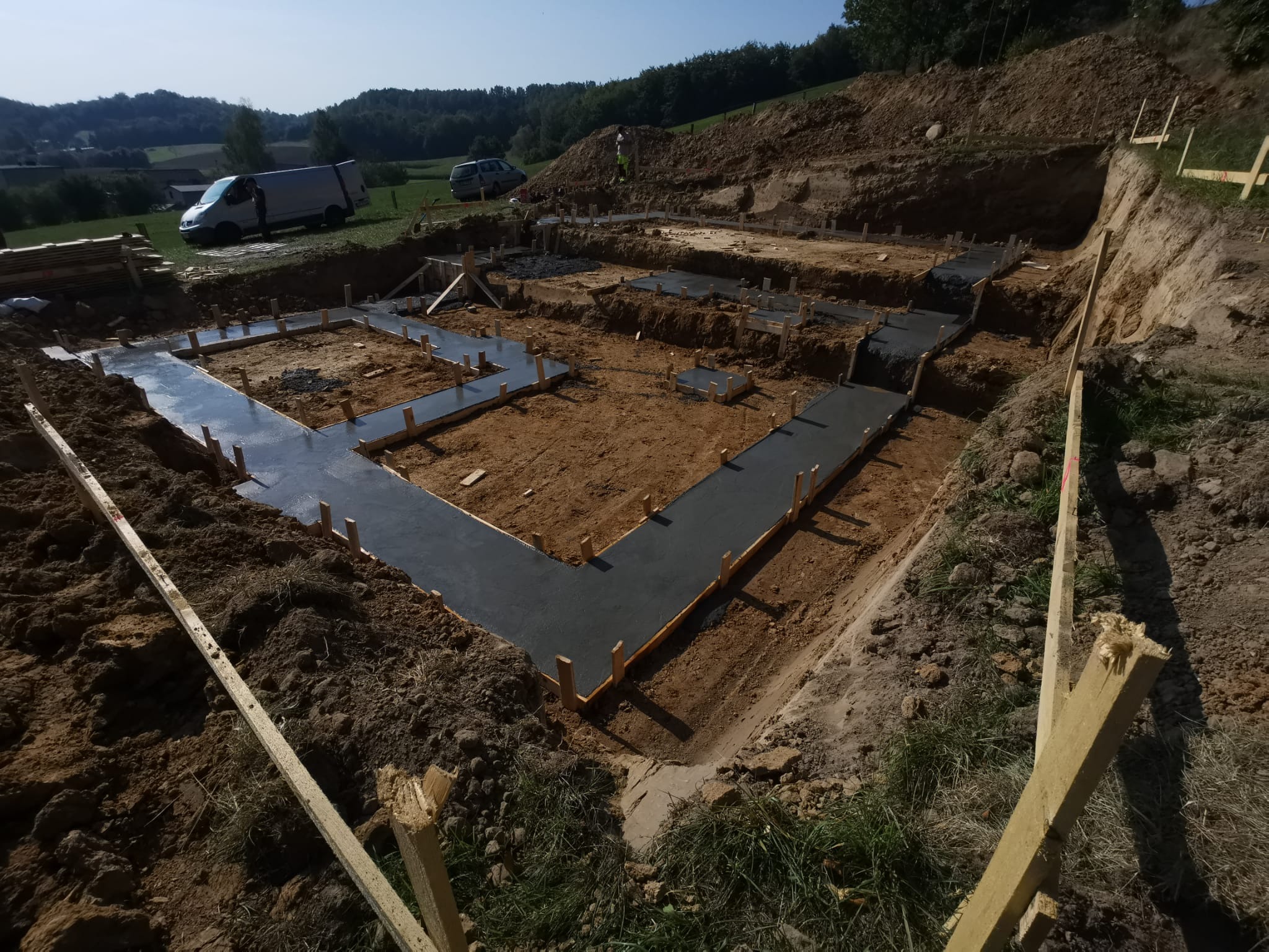 Wylane fundamenty pod budowę domu, widoczny szalunek i świeży beton, teren przygotowany pod dalsze prace budowlane, w tle krajobraz zalesiony i zaparkowane samochody.