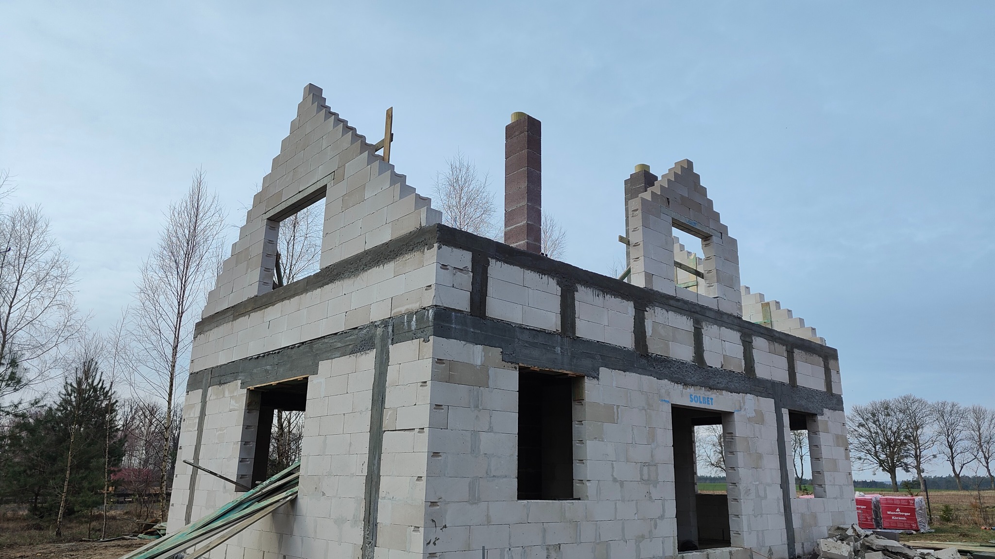 Surowy stan domu murowanego z betonu komórkowego Solbet, widoczne otwory okienne, komin z cegły klinkierowej i częściowo wykonane szczyty dachu na tle zimowego krajobrazu.