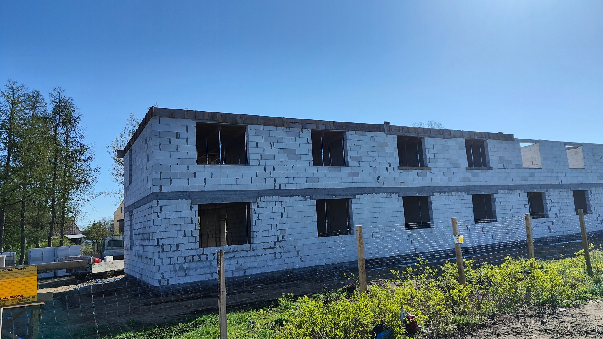 Dwukondygnacyjny budynek w trakcie budowy z białych bloczków, z widocznymi otworami okiennymi i drewnianą konstrukcją na dachu, ogrodzony siatką.