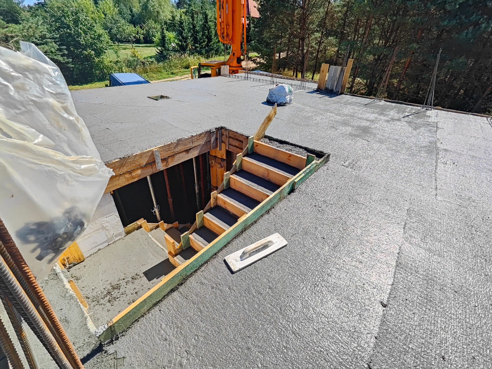 Świeżo wylany beton na stropie z przygotowanym otworem na schody, widoczna szalunkowa konstrukcja i kielnia murarska obok. W tle las i pompa do betonu.