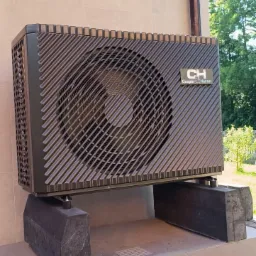 Zewnętrzna jednostka pompy ciepła Cooper&Hunter zamontowana na betonowych blokach przy jasnej ścianie budynku z widoczną rynną spustową.