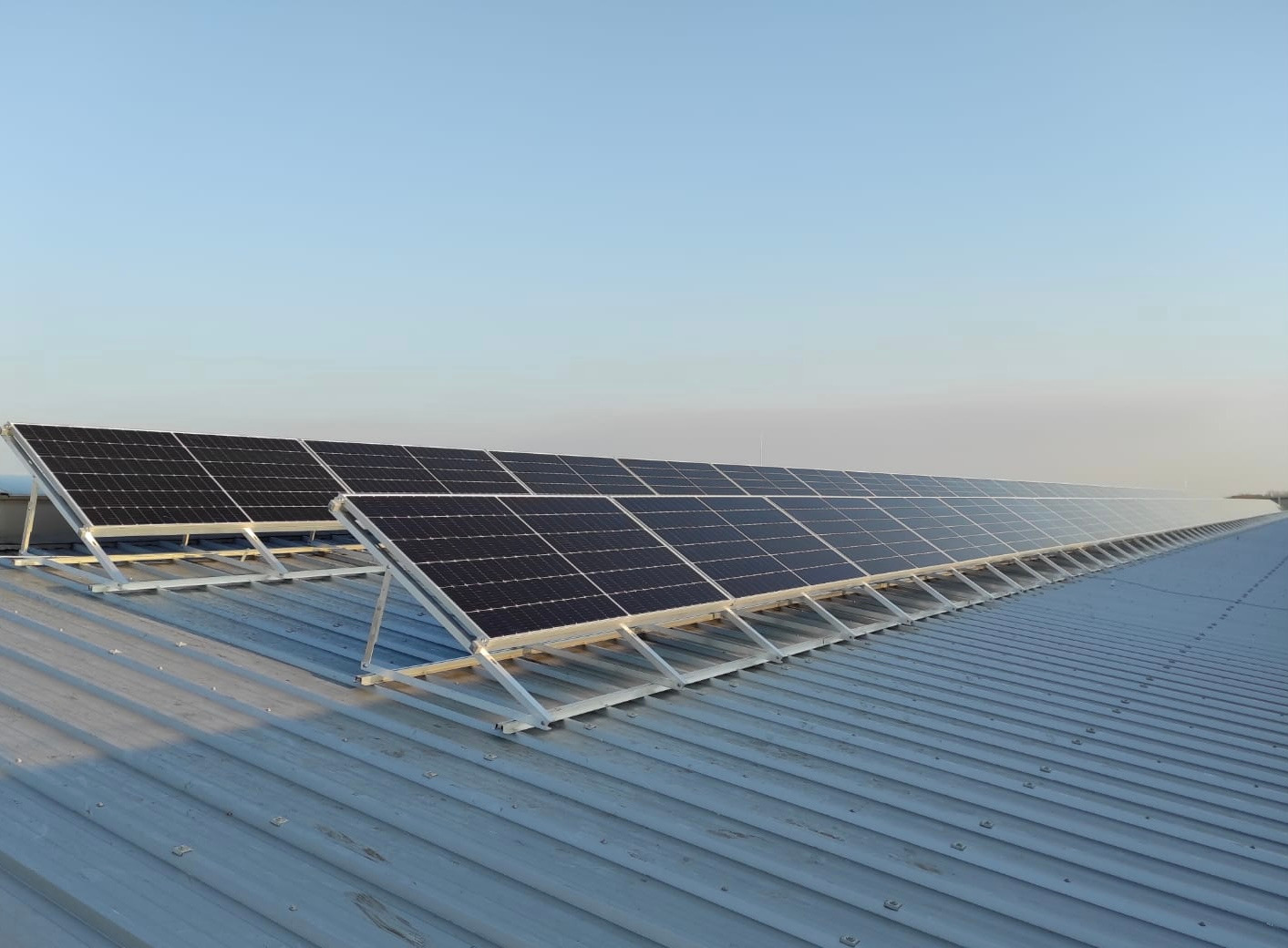 Rząd paneli fotowoltaicznych bifacial na konstrukcji wsporczej, zamontowanych na blaszanym dachu budynku przemysłowego w słoneczny dzień.