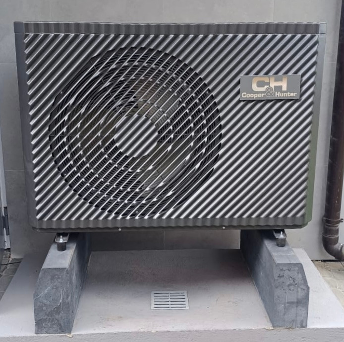 Szara zewnętrzna jednostka klimatyzacji Cooper&Hunter zamontowana na betonowych wspornikach, widoczna kratka wentylacyjna i rura spustowa po prawej stronie.