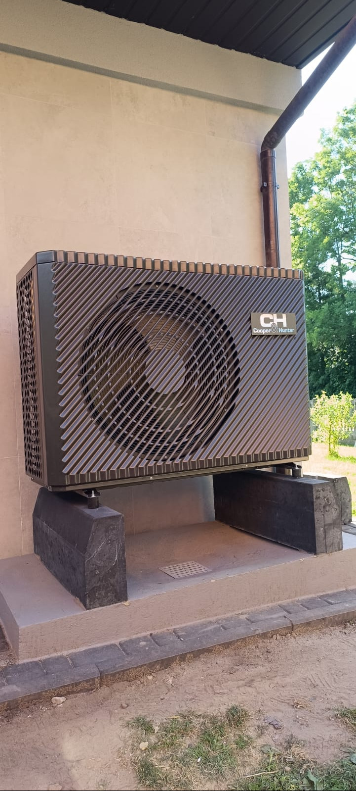 Zewnętrzna jednostka pompy ciepła Cooper&Hunter zamontowana na betonowych blokach przy jasnej ścianie budynku z widoczną rynną spustową.