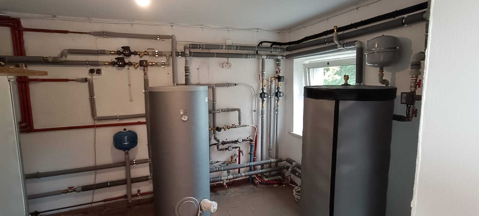 Dwa zbiorniki na wodę w kotłowni, z licznymi rurami i zaworami, widok na okno w tle.