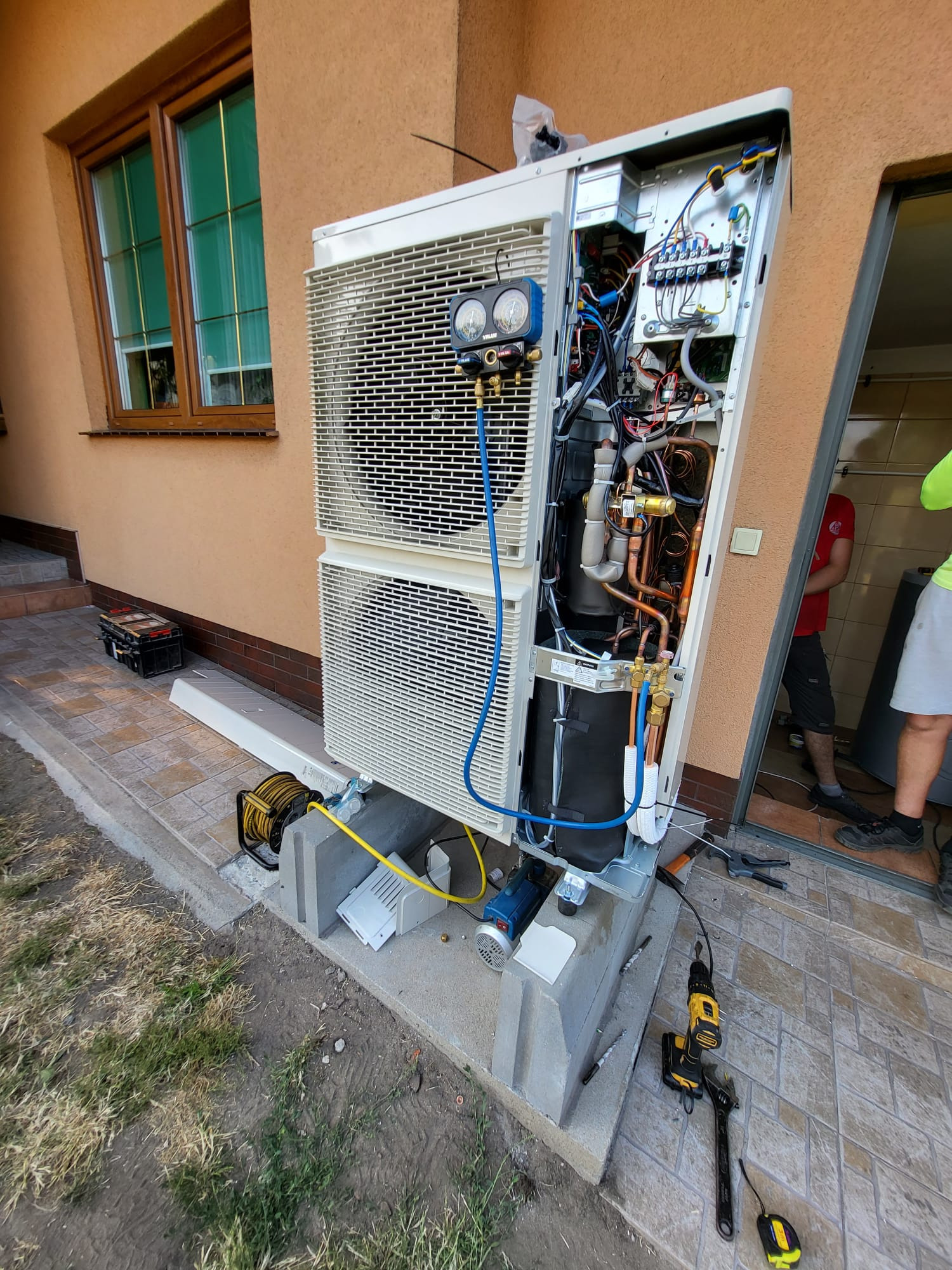 Montaż zewnętrznej jednostki pompy ciepła przy budynku, widoczne podłączenia, manometry, narzędzia pracy i częściowo sylwetka instalatora w drzwiach.