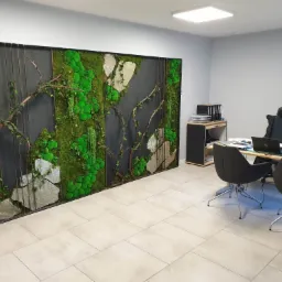 Panel dekoracyjny z mchu, kamieni i gałęzi na ścianie biura z szarymi ścianami i jasnymi płytkami podłogowymi, widoczne biurko i krzesła.