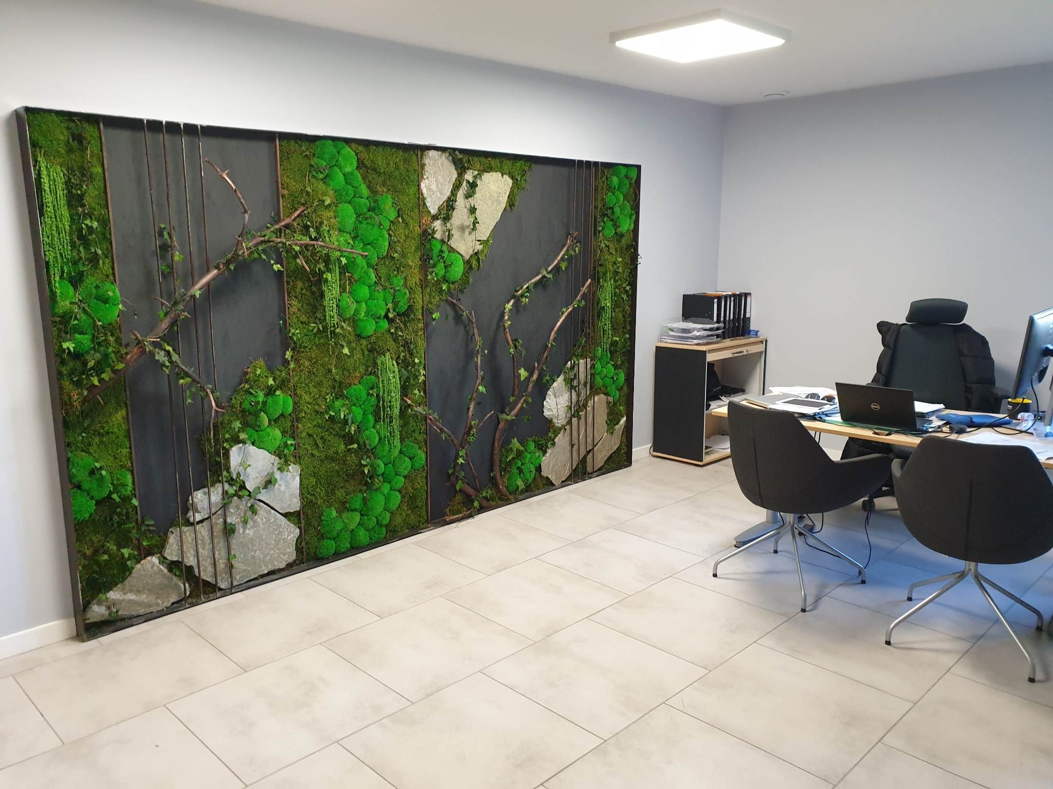 Panel dekoracyjny z mchu, kamieni i gałęzi na ścianie biura z szarymi ścianami i jasnymi płytkami podłogowymi, widoczne biurko i krzesła.